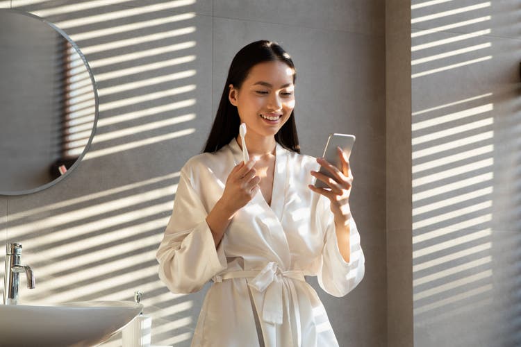 Cheerful long-haired millennial korean lady in silk bathrobe using mobile phone while doing morning routine in bathroom, brushing teeth, looking at smartphone screen and smiling, sun flare